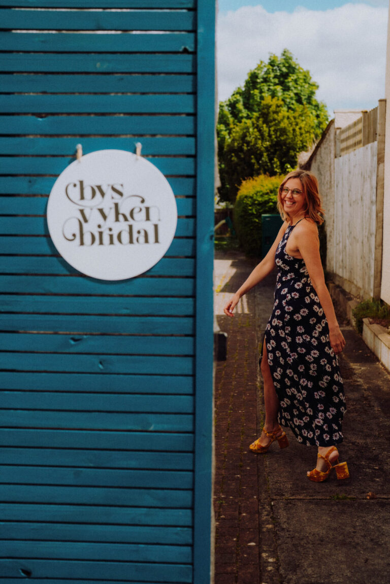 Photograph of Zoe, owner of Bys Vyken Bridal, smiling as she looks at the camera over her shoulder and walks past a sign showing the Bys Vyken Bridal logo